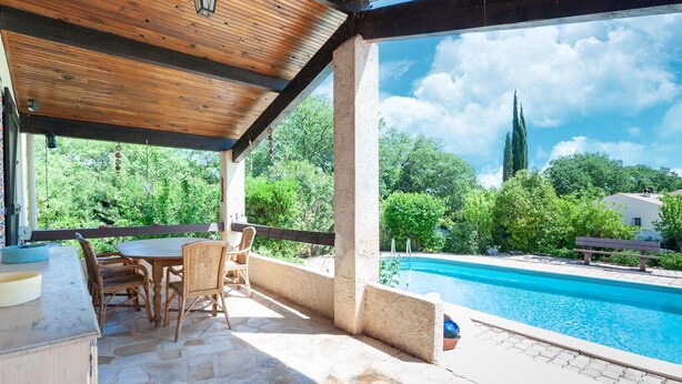 Un patio al aire libre con piso de piedra, una mesa de madera y sillas de mimbre. Se ve una piscina al fondo, con vistas a árboles y un edificio.