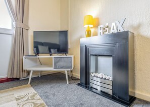 Una sala de estar moderna con un televisor de pantalla plana sobre un soporte blanco, una chimenea con un letrero 'FLAX' encima y una lámpara amarilla.