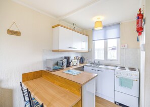Een kleine keuken met witte kastjes, een houten tafel, een spoelbak, een fornuis en een raam.
