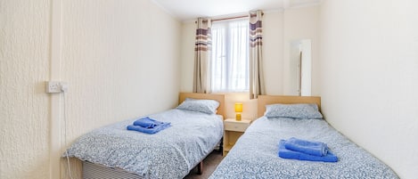 Una habitación pequeña y limpia con dos camas, una mesita de noche, una lámpara, una ventana con cortinas y un enchufe.