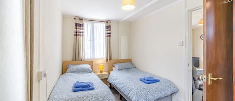 Quarto compacto com duas camas de solteiro, mesa de cabeceira com abajur, janela com cortinas e porta que dá para outro cômodo.
