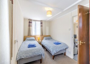 Een kleine slaapkamer met twee eenpersoonsbedden, een nachtkastje met een lamp, een raam met gordijnen en een deur naar een andere kamer.