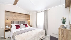 Een moderne slaapkamer met een houten hoofdbord, een groot bed met veel kussens, een nachtkastje met een lamp en een bijzettafeltje met een fotolijst.