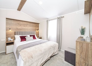 A modern bedroom with a wooden headboard, a large bed with multiple pillows, a bedside table with a lamp, and a nightstand with a picture frame.