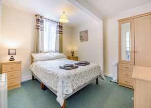 Une chambre à coucher comprenant un lit, des tables de chevet, un radiateur, une armoire et une fenêtre avec des rideaux.
