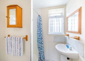 Une salle de bain avec un meuble en bois, un porte-serviettes orné d’une serviette à motifs, un rideau de douche, une fenêtre avec des rideaux en dentelle et un lavabo avec un flacon de savon liquide.