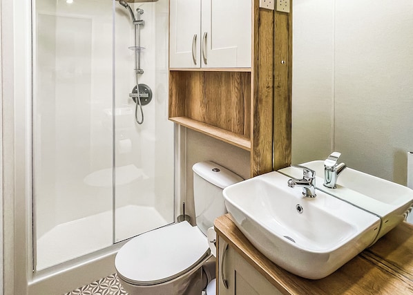 A bathroom with a glass shower enclosure, a white toilet, a wooden vanity with a sink, and a mirror. - The Amy Johnson Lodge (Tunstall, Nr Hull)