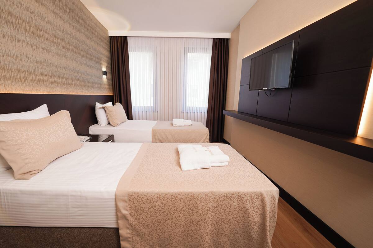 A hotel room with two beds, a flat-screen TV mounted on the wall, and a large window with curtains.