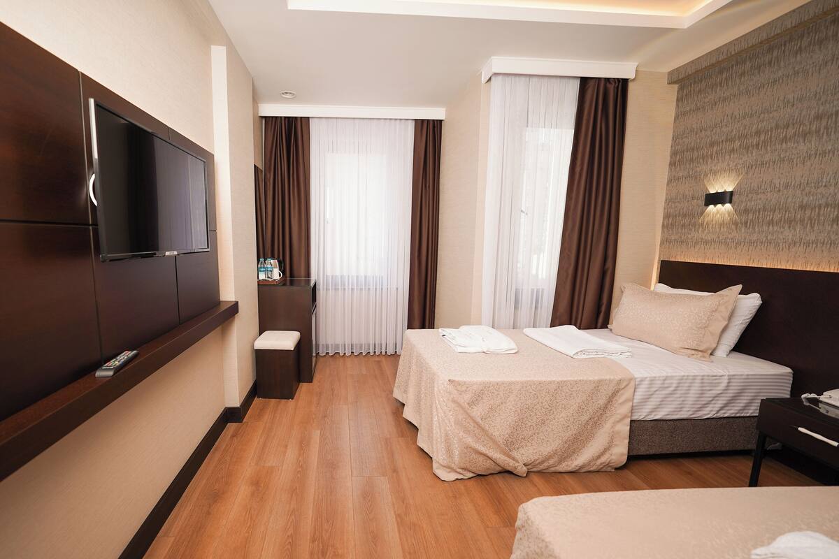 A hotel room with a flat-screen TV mounted on the wall, a bed with a headboard, a bedside table, and a small desk with a chair.