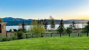 A grassy field with a wooden fence, a building, and a lake with mountains in the background. - Hilltop Home on Lake Whatcom | Views, Beach Access & Space for Ten (Bellingham)