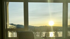 A room with a patterned armchair and a small round table, large windows with a view of a sunset over a body of water and hills. - Hilltop Home on Lake Whatcom | Views, Beach Access & Space for Ten (Bellingham)