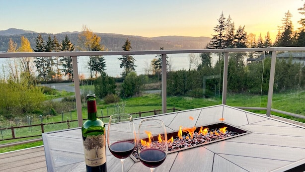 A table with a bottle of wine and two glasses on a balcony overlooking a lake and forest. - Hilltop Home on Lake Whatcom | Views, Beach Access & Space for Ten (Bellingham)