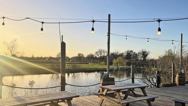 A wooden deck with picnic tables overlooking a calm body of water, string lights, and a clear sky. - 1 bedroom accommodation in Old Buckenham (Old Buckenham)