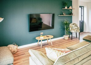 A living room with a flat-screen TV mounted on the wall, a round wooden coffee table, a sofa, a chair, and a potted plant.