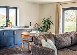 Eine moderne Küche mit dunklen Schränken, einem Esstisch und Stühlen. Ein großes Fenster mit Vorhängen, eine Pflanze und ein Sofa gehören zur Wohnbereich.