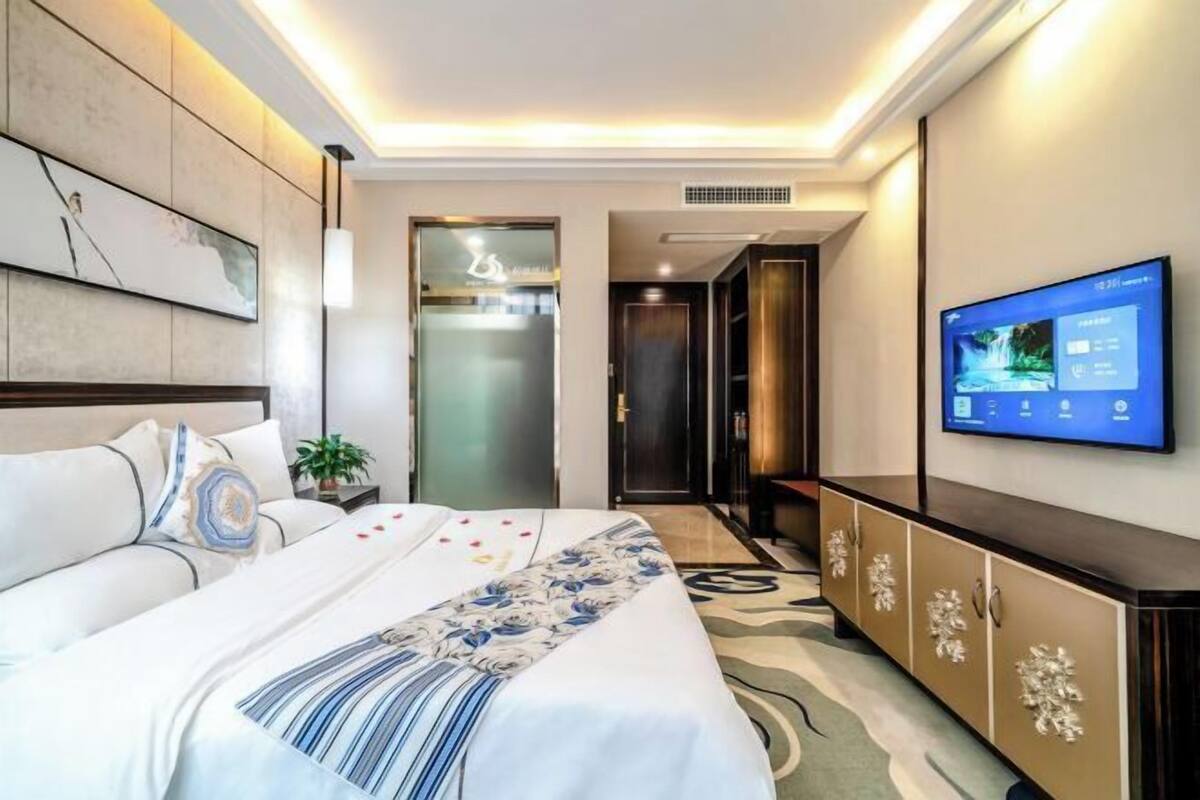 A modern hotel room with a large bed, a flat-screen TV mounted on the wall, and a minibar.