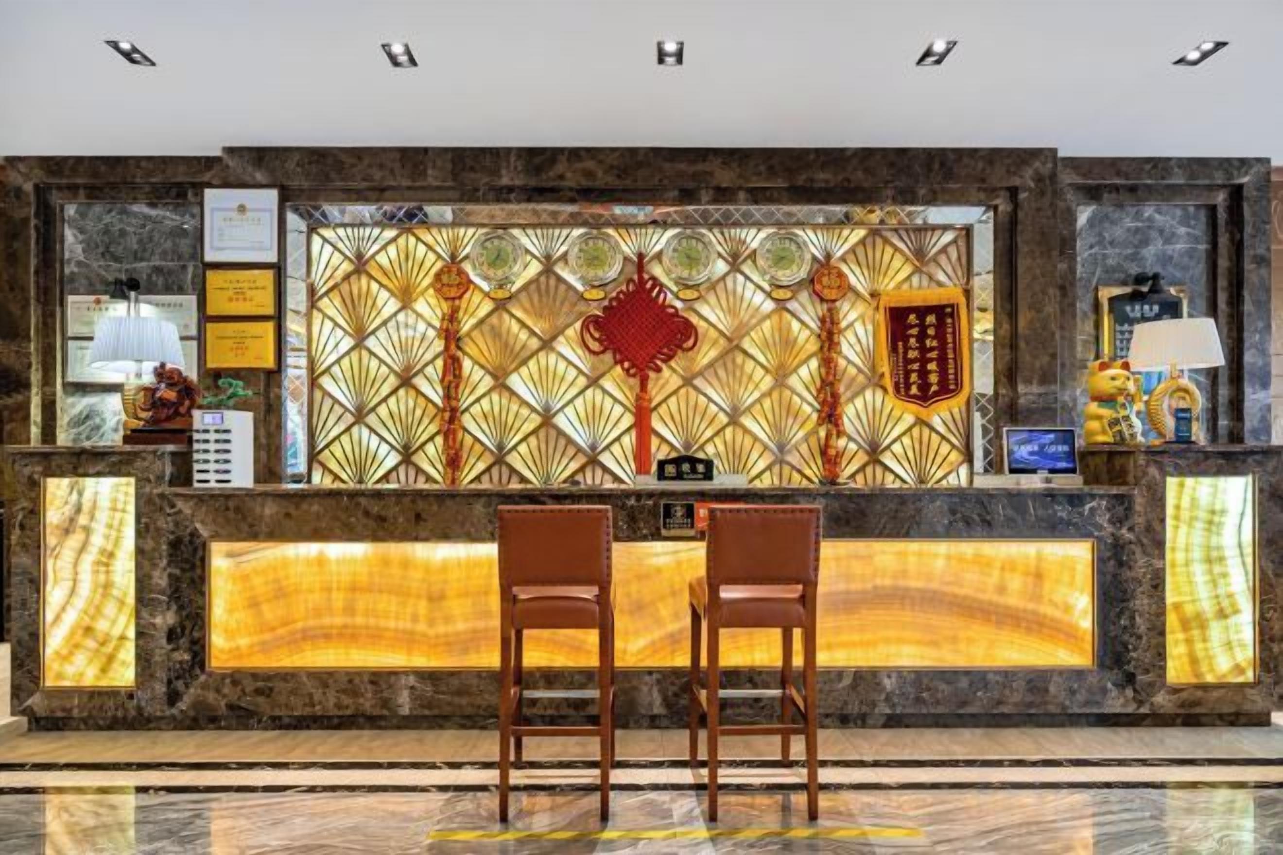 Recepção de hotel com balcão de mármore, duas cadeiras de madeira e parede decorativa com padrões geométricos.