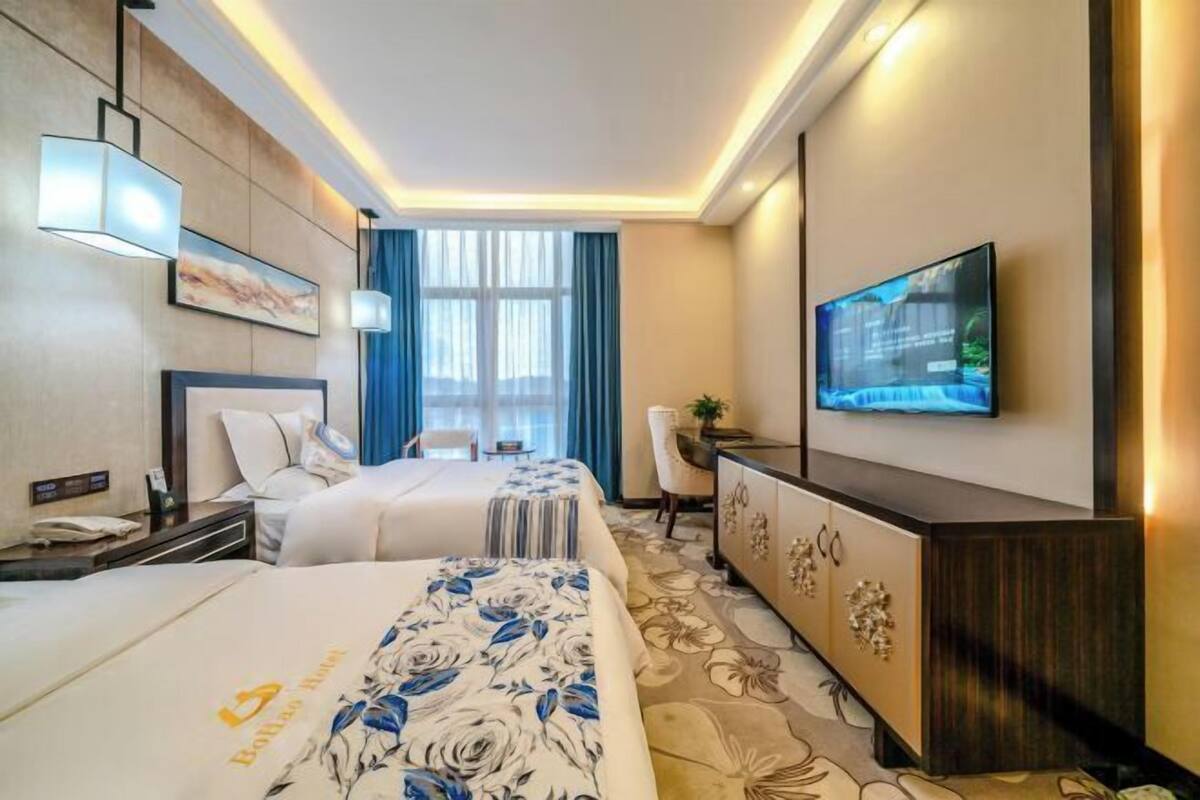 A hotel room with two beds, a desk, a TV, and a large window with curtains.