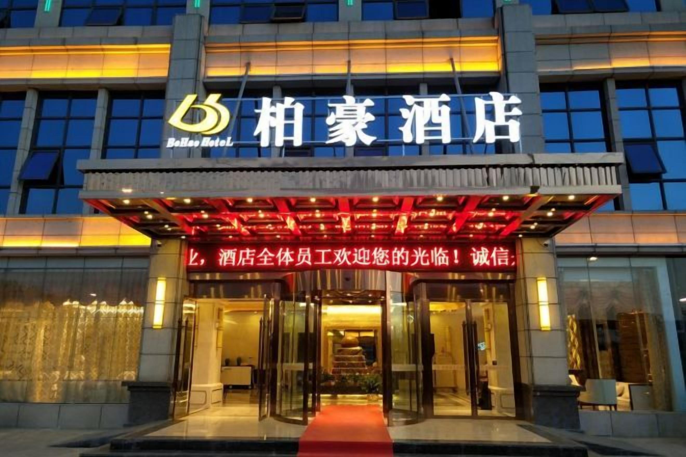 A hotel entrance with a red carpet leading to the doors, a signboard with hotel name, and a modern facade with large windows.