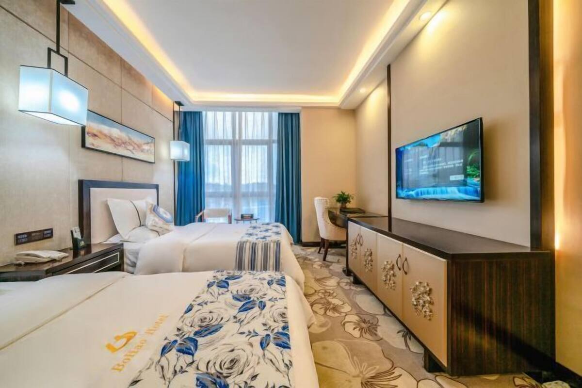 A hotel room with two beds, a TV, a desk, and a large window with curtains.