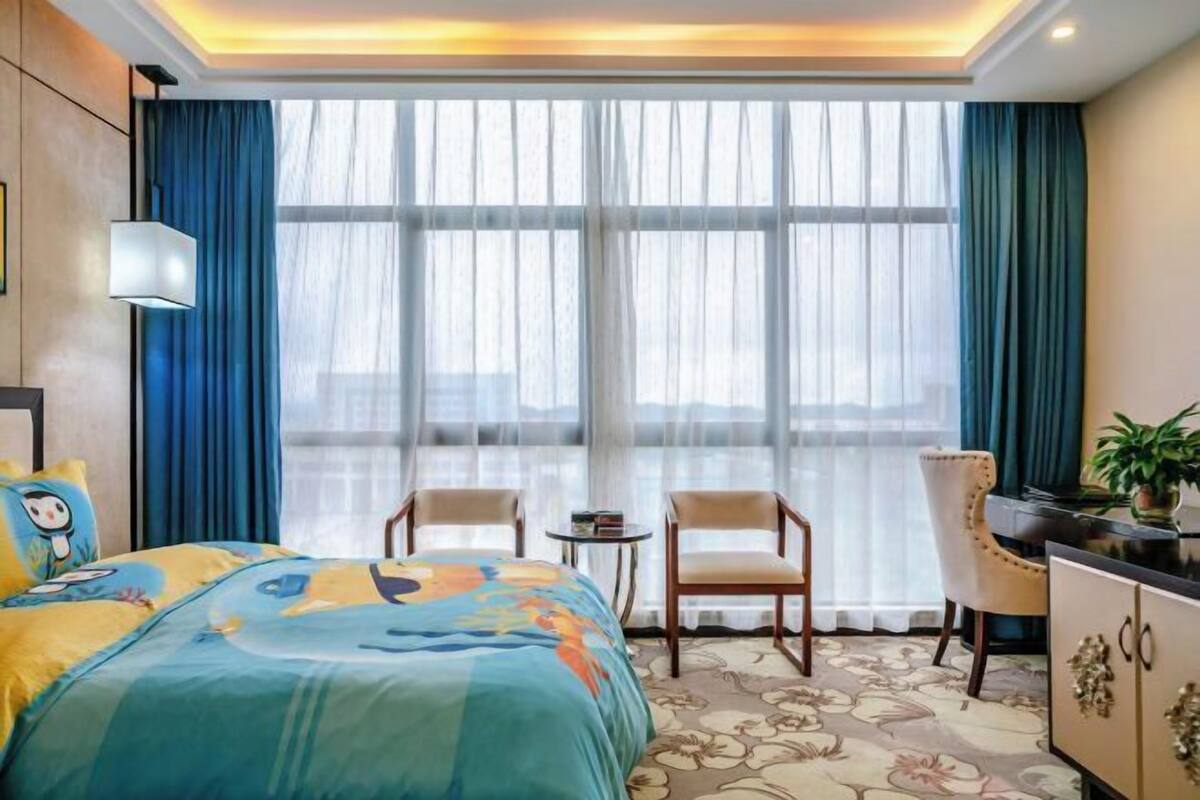 A hotel room with a bed, two chairs, a small table, a cabinet, and a large window with curtains.