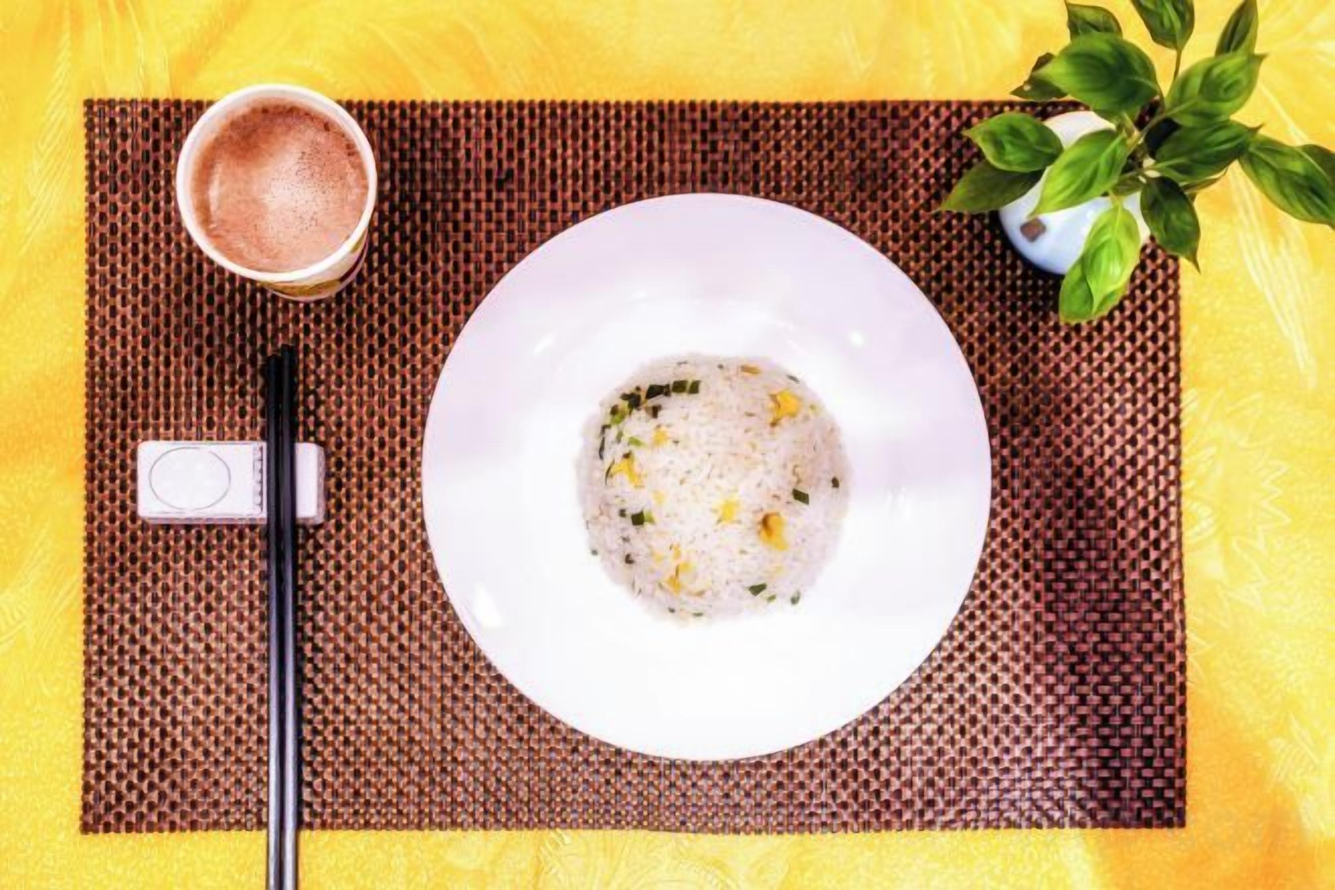A white plate with a dish, a pair of chopsticks, a cup of a beverage, and a plant on a yellow surface.