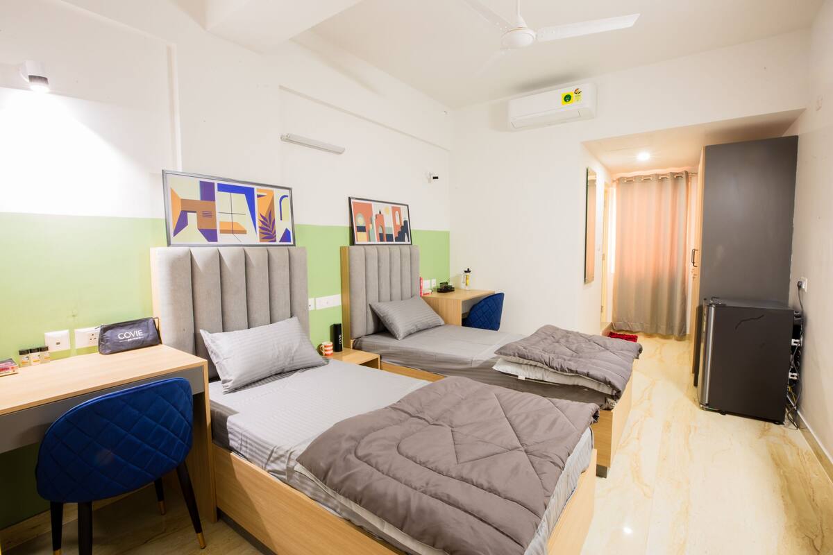 A hotel room with two beds, a desk, a chair, a refrigerator, and artwork on the walls.