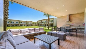 A spacious outdoor seating area with a long sofa, a dining table, and chairs, overlooking a well-maintained lawn and a building complex.