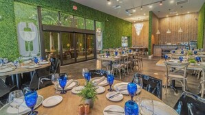 A restaurant with round tables set with blue glassware, white plates, and silverware. There are chairs around the tables, and a potted plant on one table. The walls are green with a leafy pattern, and there is a large window in the background.