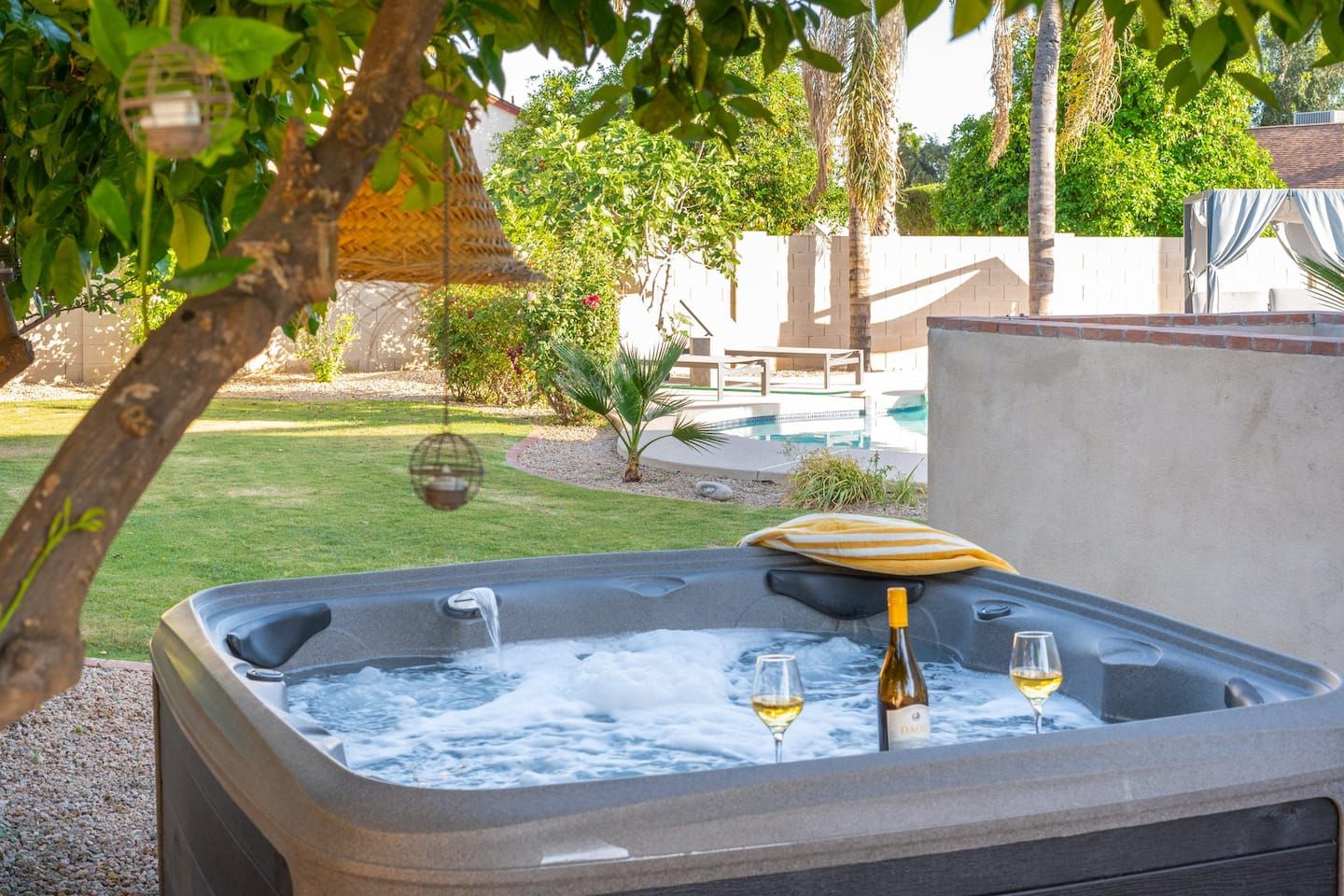 A hot tub with two glasses of wine and a bottle, surrounded by a garden and a pool.