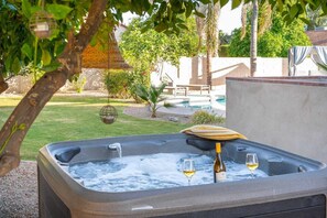A hot tub with two glasses of wine and a bottle, surrounded by a garden and a pool.