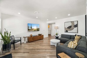 A modern living room with a grey sofa, a wooden coffee table, and a black bench. There is a wooden cabinet with decorative items and a painting of a desert landscape.