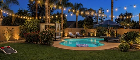 Un patio trasero con piscina, luces colgantes, palmeras y mobiliario exterior.