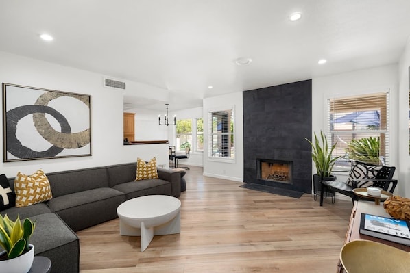 A modern living room with a fireplace, a sofa, a coffee table, and a dining area in the background.