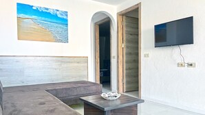 A modern living room with a curved sofa, a small table with a decorative bowl, a flat-screen TV mounted on the wall, and a wooden door leading to another room.