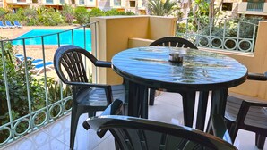 A balcony with a round table and chairs, overlooking a swimming pool and a resort building with multiple balconies and palm trees.