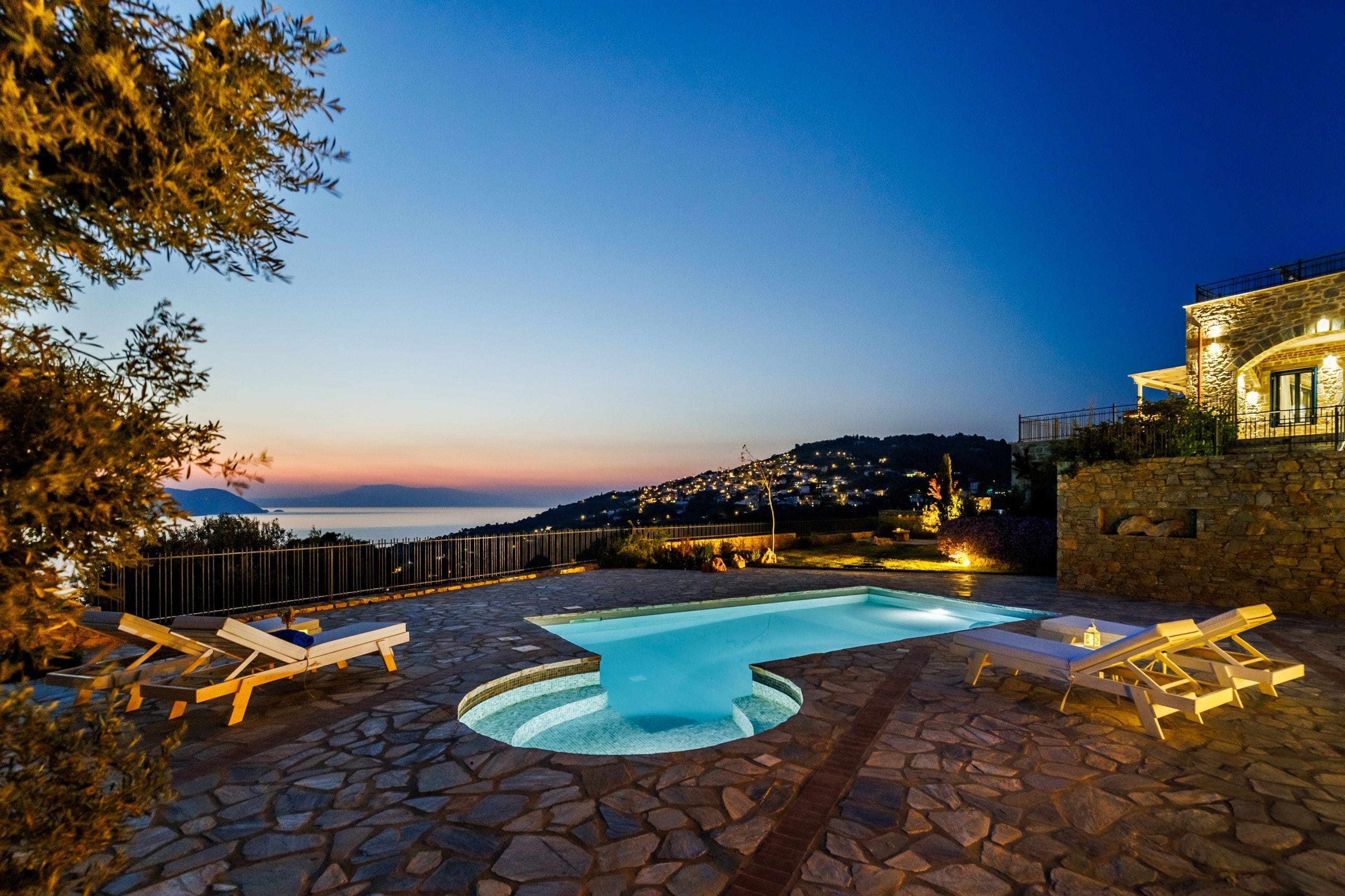 A stone pool with lounge chairs, a stone deck, and a view of a hillside at dusk.