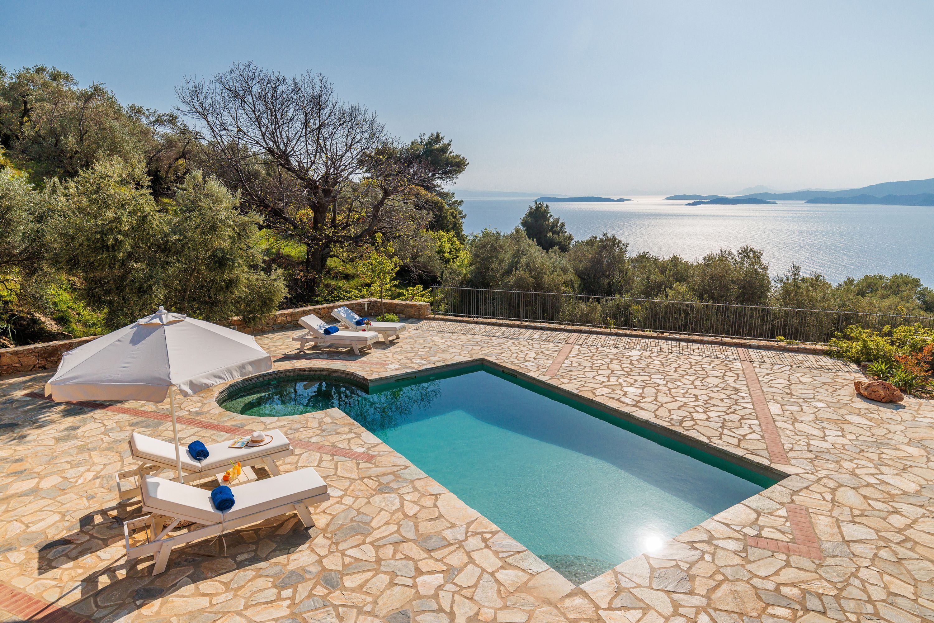 A rectangular swimming pool with stone tiles, surrounded by lounge chairs and an umbrella, overlooking a body of water.