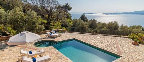 A rectangular swimming pool with stone tiles, surrounded by lounge chairs and an umbrella, overlooking a body of water.