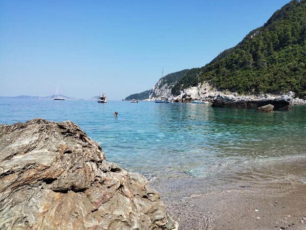 A rocky shoreline with clear water, boats, and a mountainous backdrop.