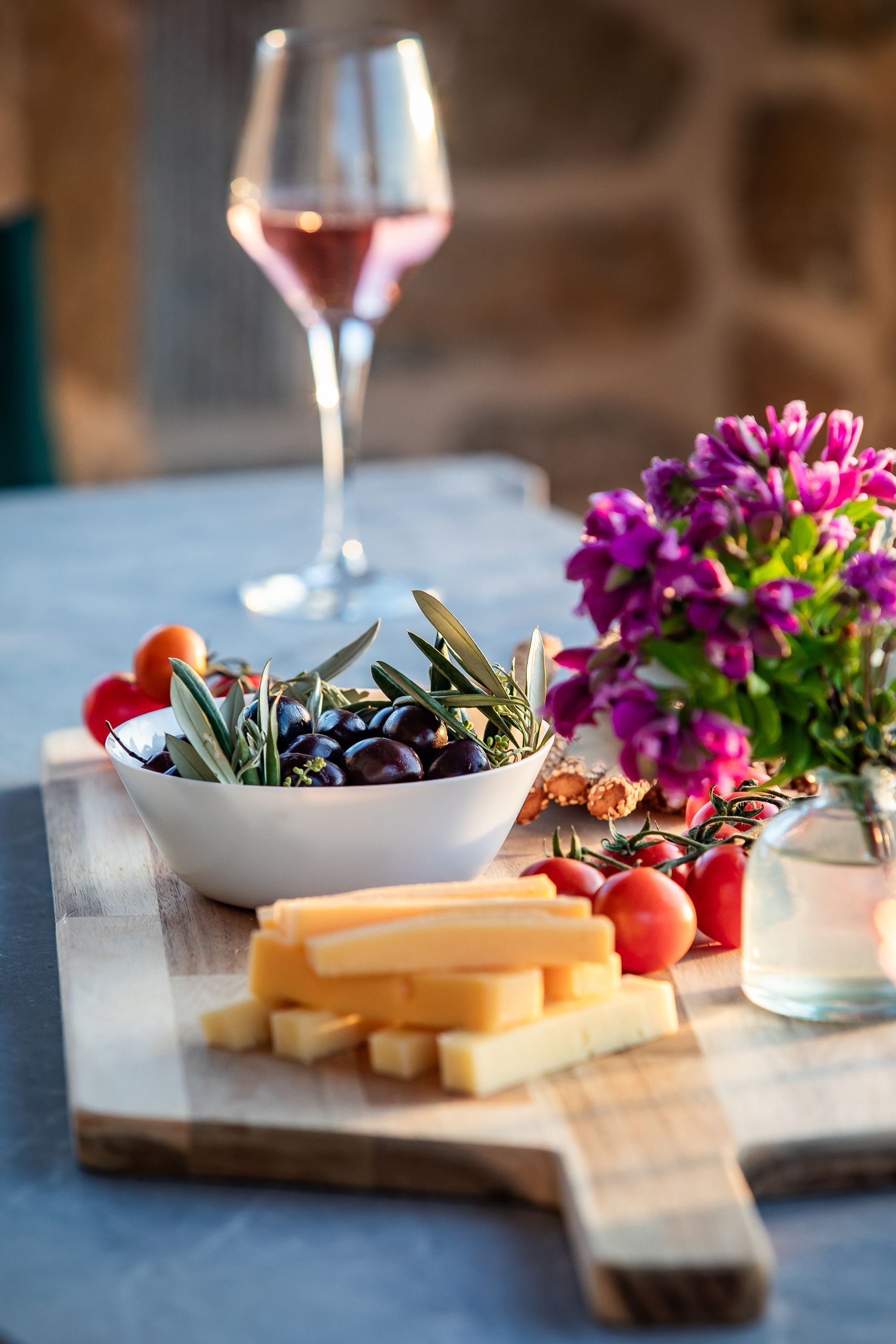 A table with a bowl of olives, tomatoes, and cheese, a glass of red wine, and a vase with purple flowers.