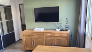 A modern living room with a wooden dining table, a flat-screen TV mounted on the wall, and a large window with curtains. - Appartement Dans une Résidence ART Deco!! au Bord de la Plage (La Ciotat)