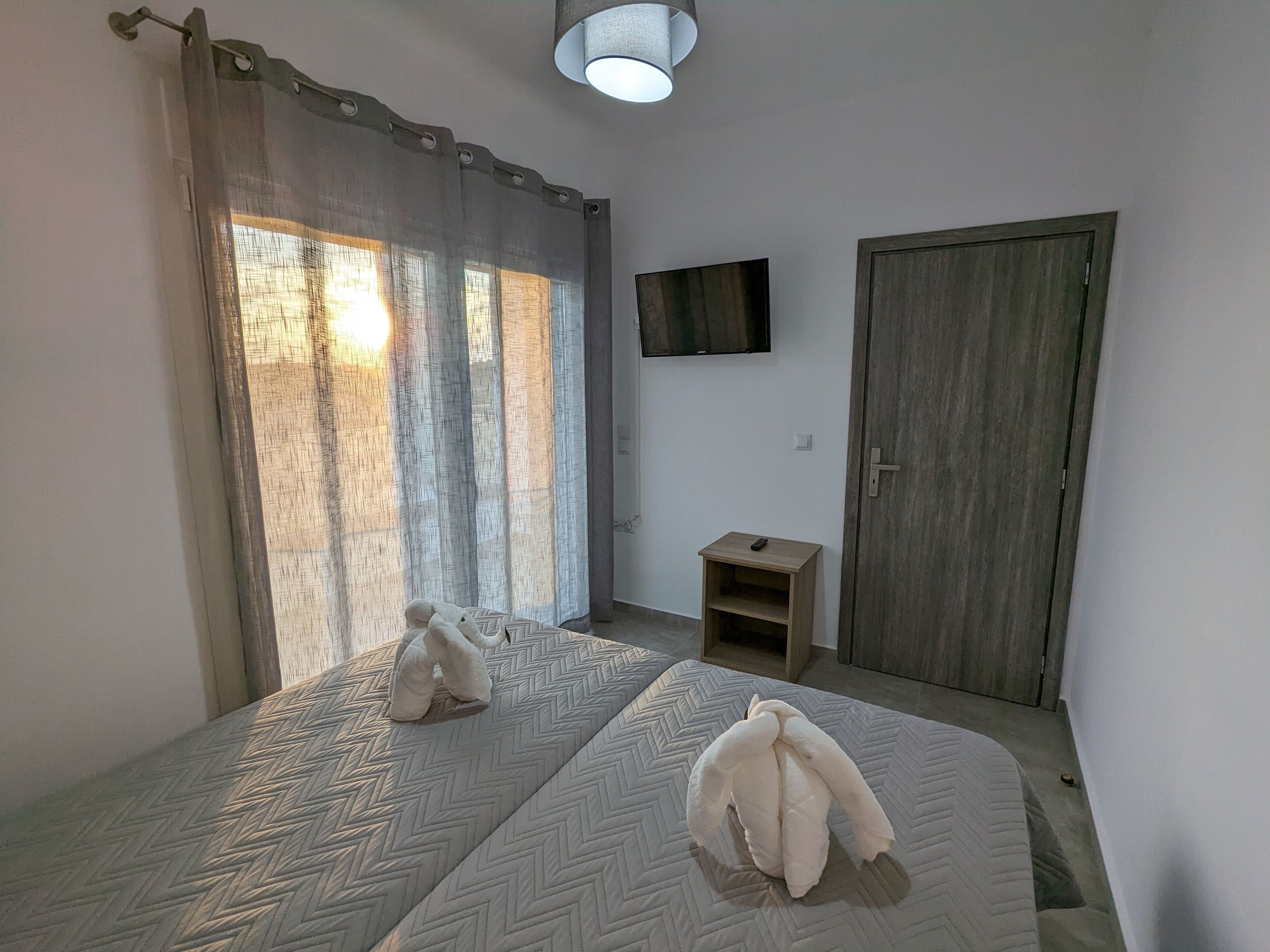 A room with two beds, a TV on the wall, a wooden door, and a small table with a remote control.