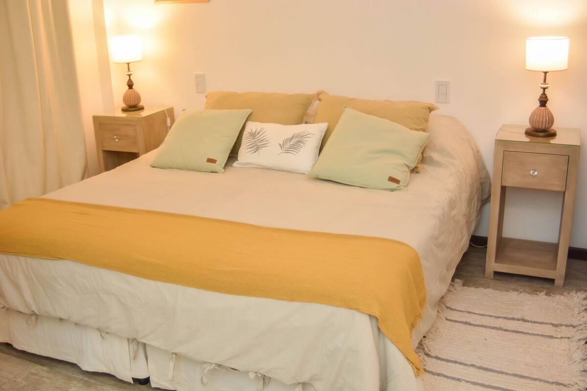 Una cama bien tendida con una colcha amarilla y blanca, dos mesitas de noche con lámparas y una alfombra a rayas en el suelo.