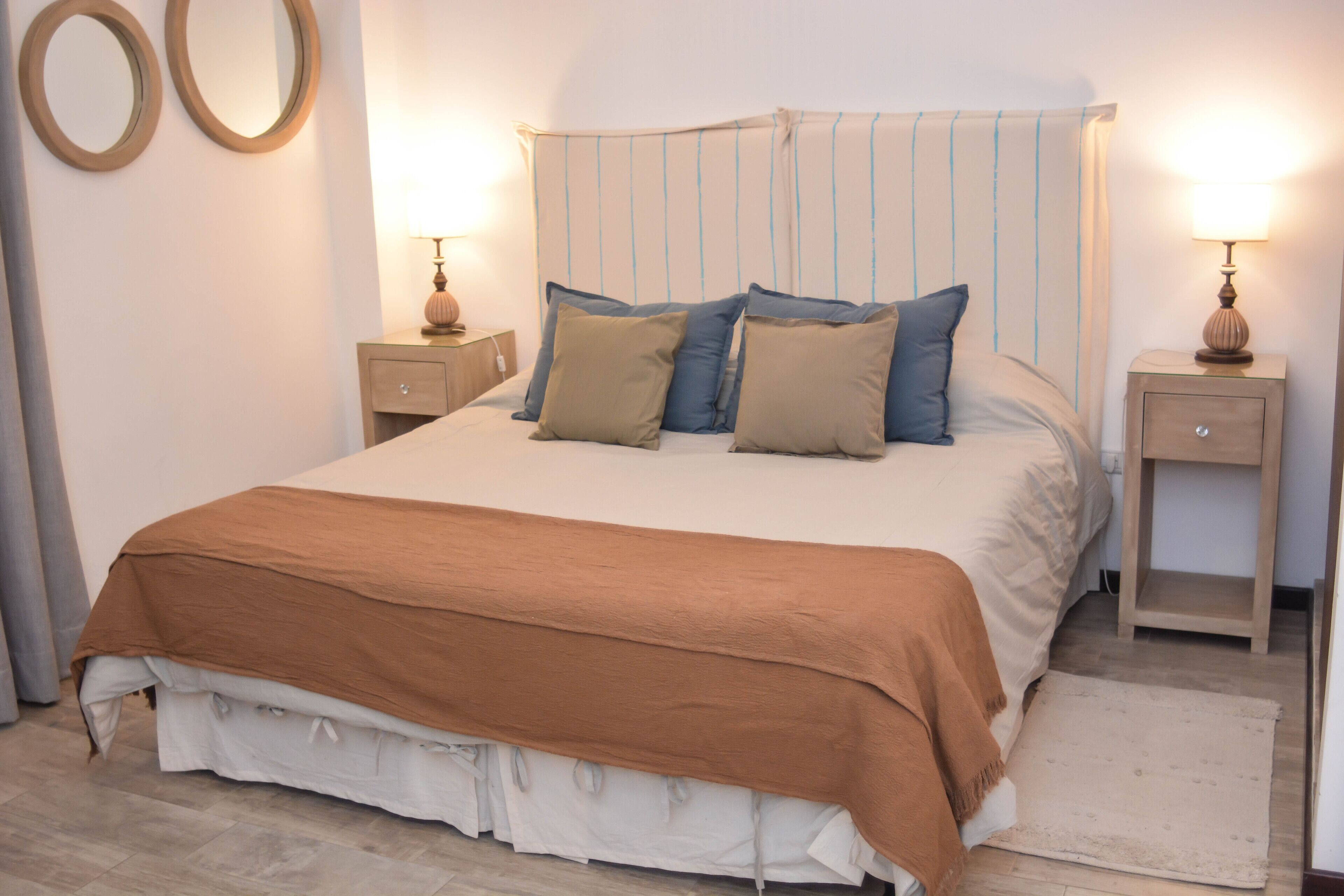 Una cama bien hecha con una colcha marrón y almohadas, dos mesitas de noche con lámparas, y un espejo en la pared.