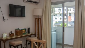 A hotel room with a TV mounted on the wall, a desk with a lamp, a chair, and a view of the outside balcony.