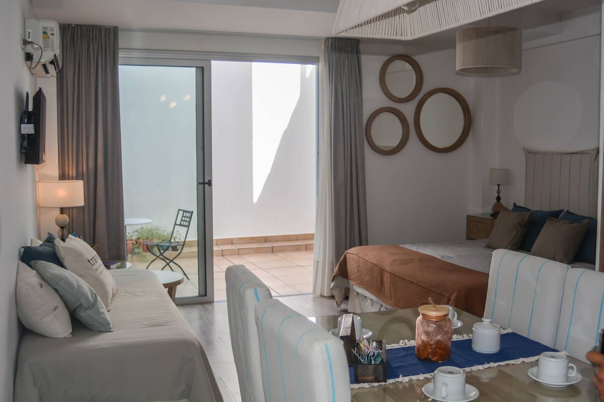 Habitación de hotel con cama, sofá, mesa de comedor con tazas y un tarro, y vistas al exterior.