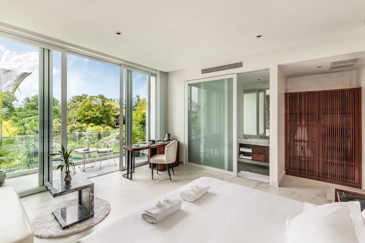 Quarto moderno com uma cama grande, uma escrivaninha com cadeira, box de chuveiro em vidro e vista para um jardim exuberante.