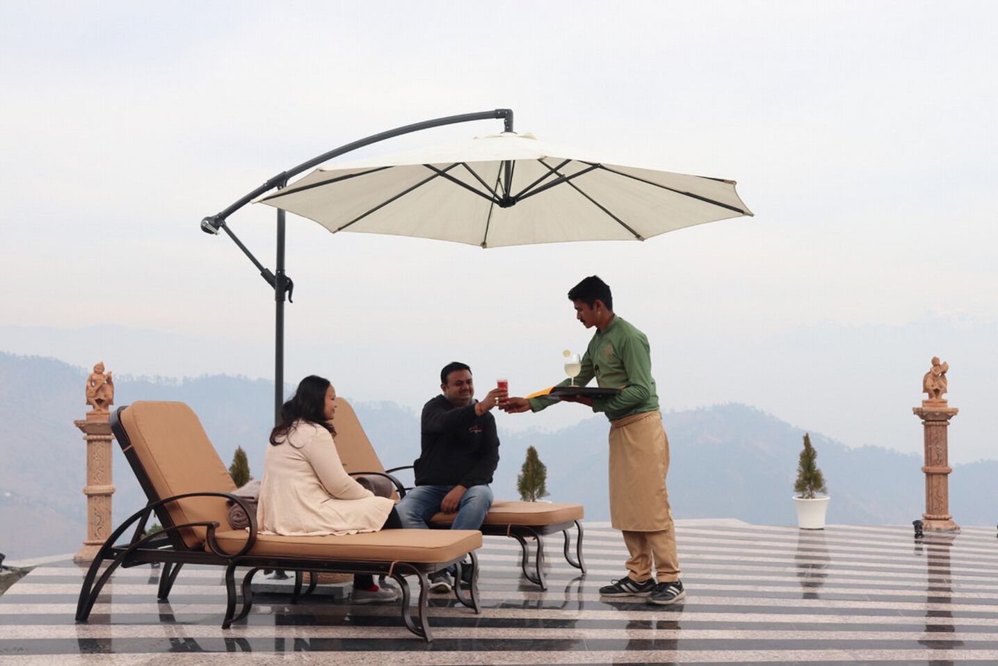 A terrace with three lounge chairs, a waiter serving a drink, and a view of the mountains.