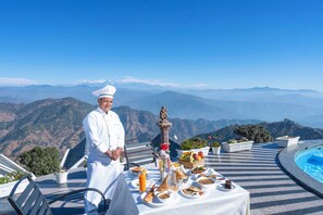Un chef se tient près d’une table où sont disposés différents plats, et on aperçoit une piscine à l’arrière-plan.